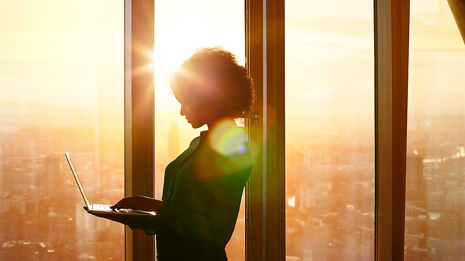Женщина, стоящая с ноутбуком на фоне заката за окнами здания