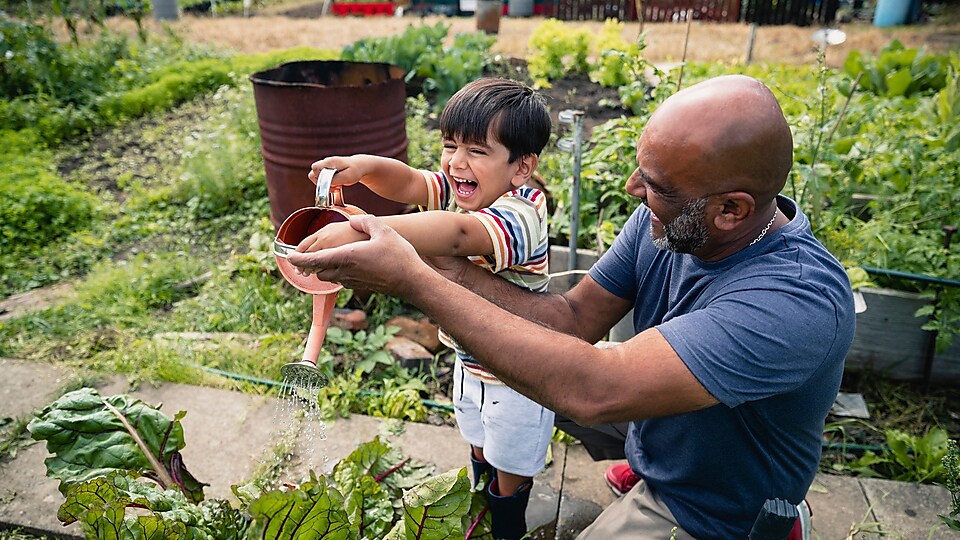 Мужчина помогает улыбающемуся ребёнку поливать растения в саду